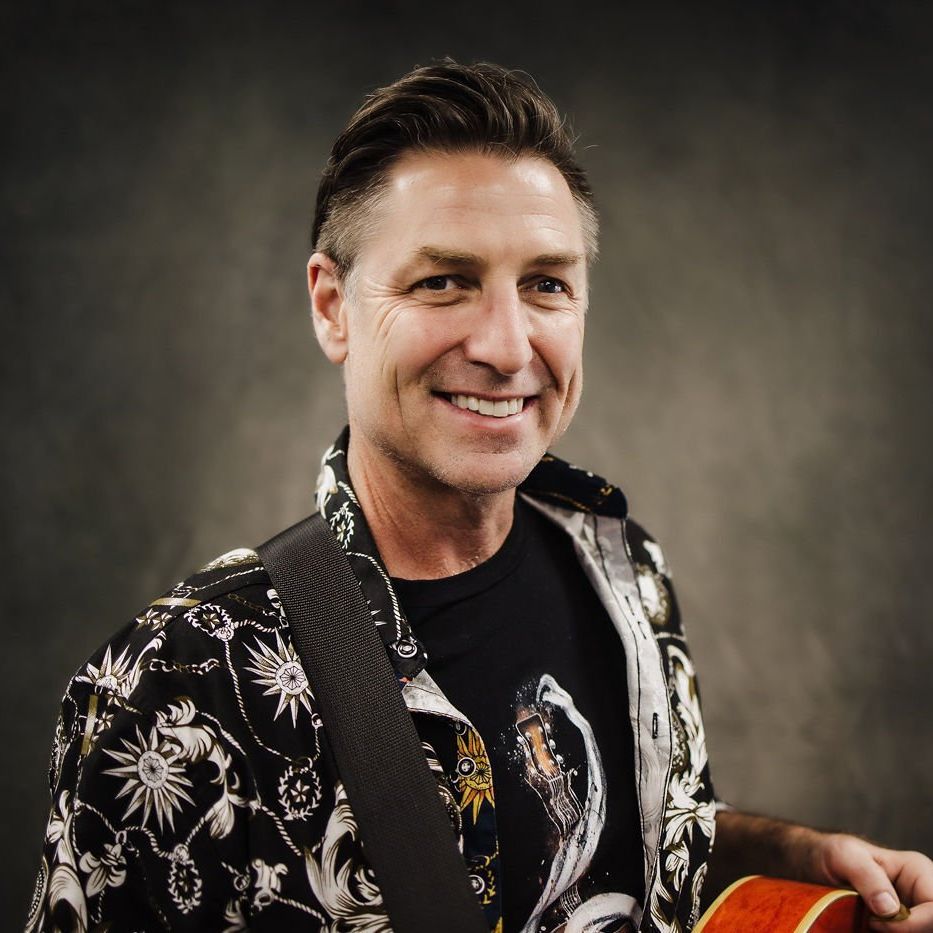 Smiling man in a black floral shirt, holding a red hat against a neutral studio backdrop