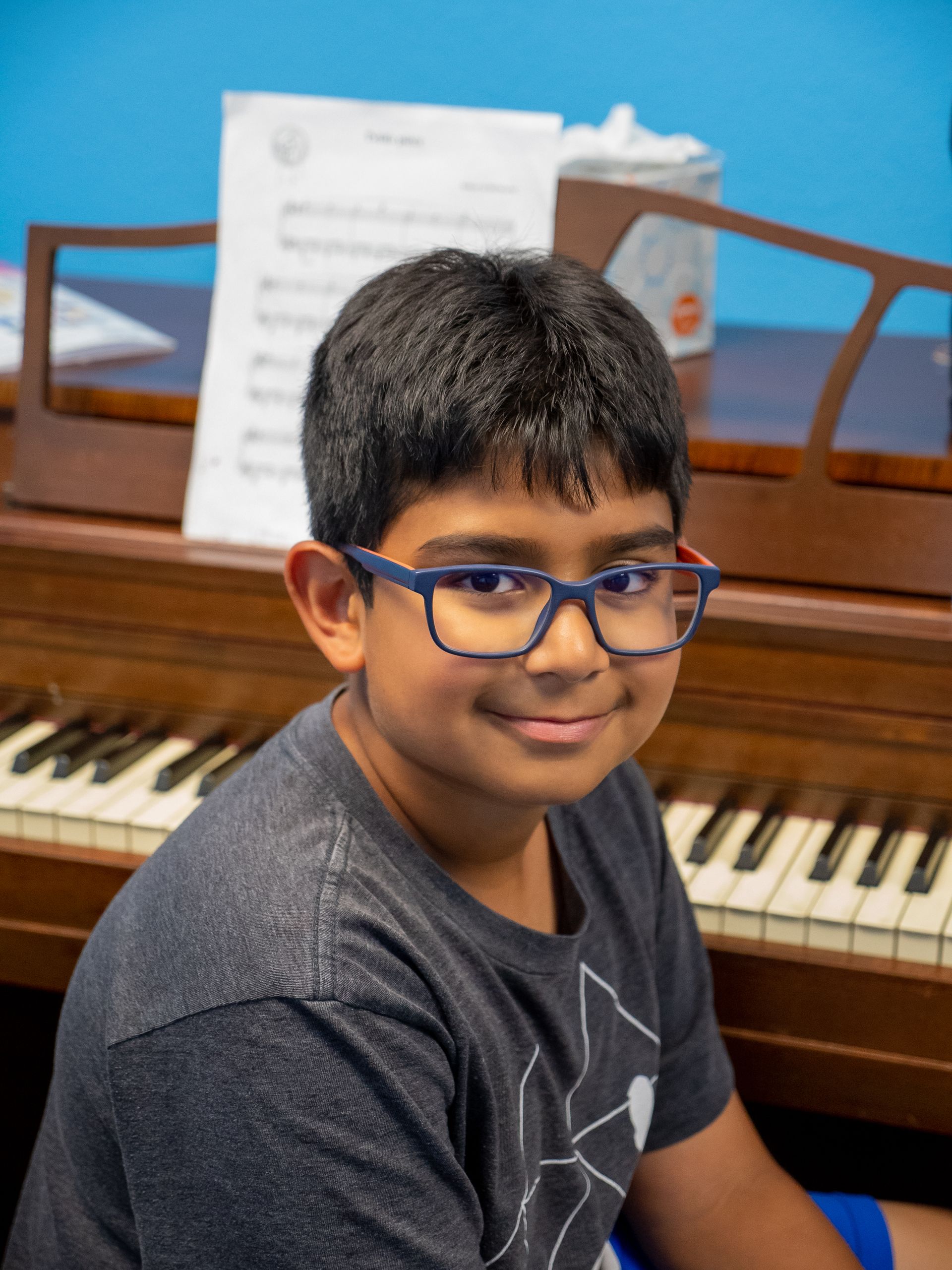Smiling child in blue glasses seated at a piano, with sheet music in the background