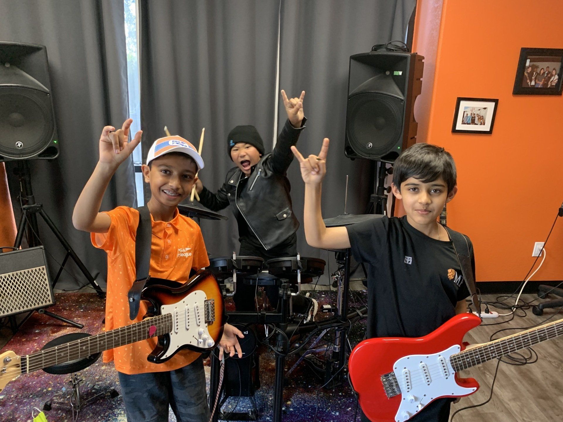 Three kids posing with electric guitars in a music studio, smiling and making rock hand signs.