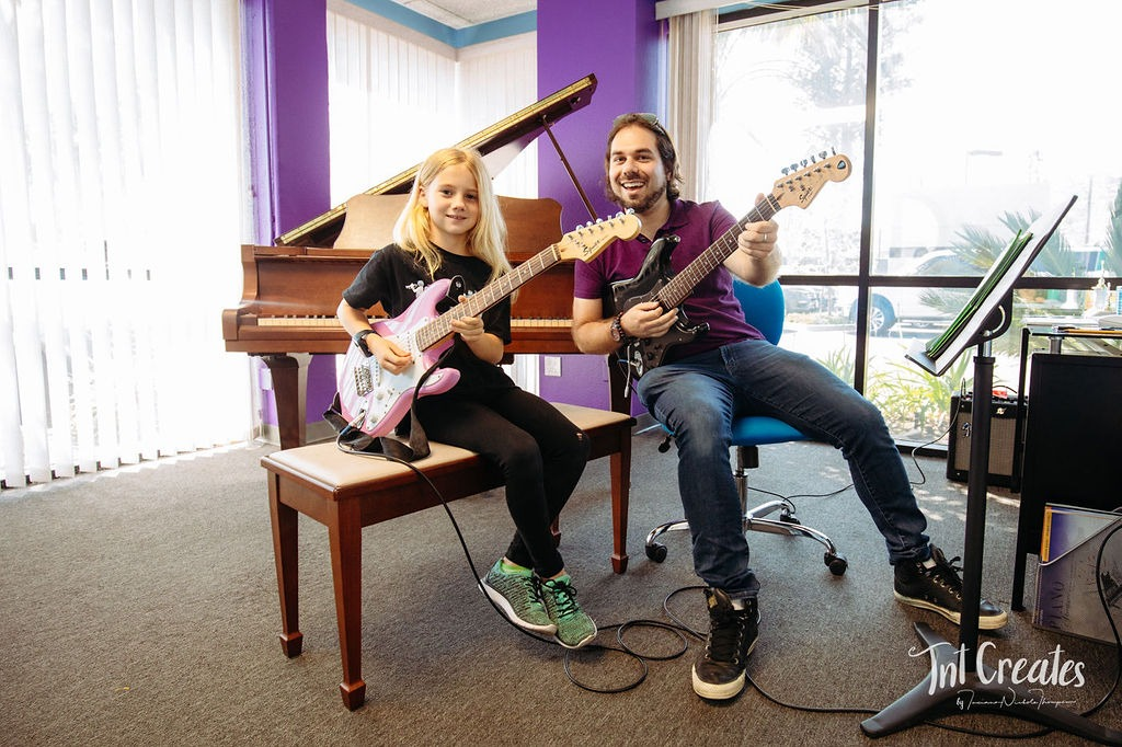 Two people sit at a piano bench playing electric guitars in a bright room with a piano behind them.