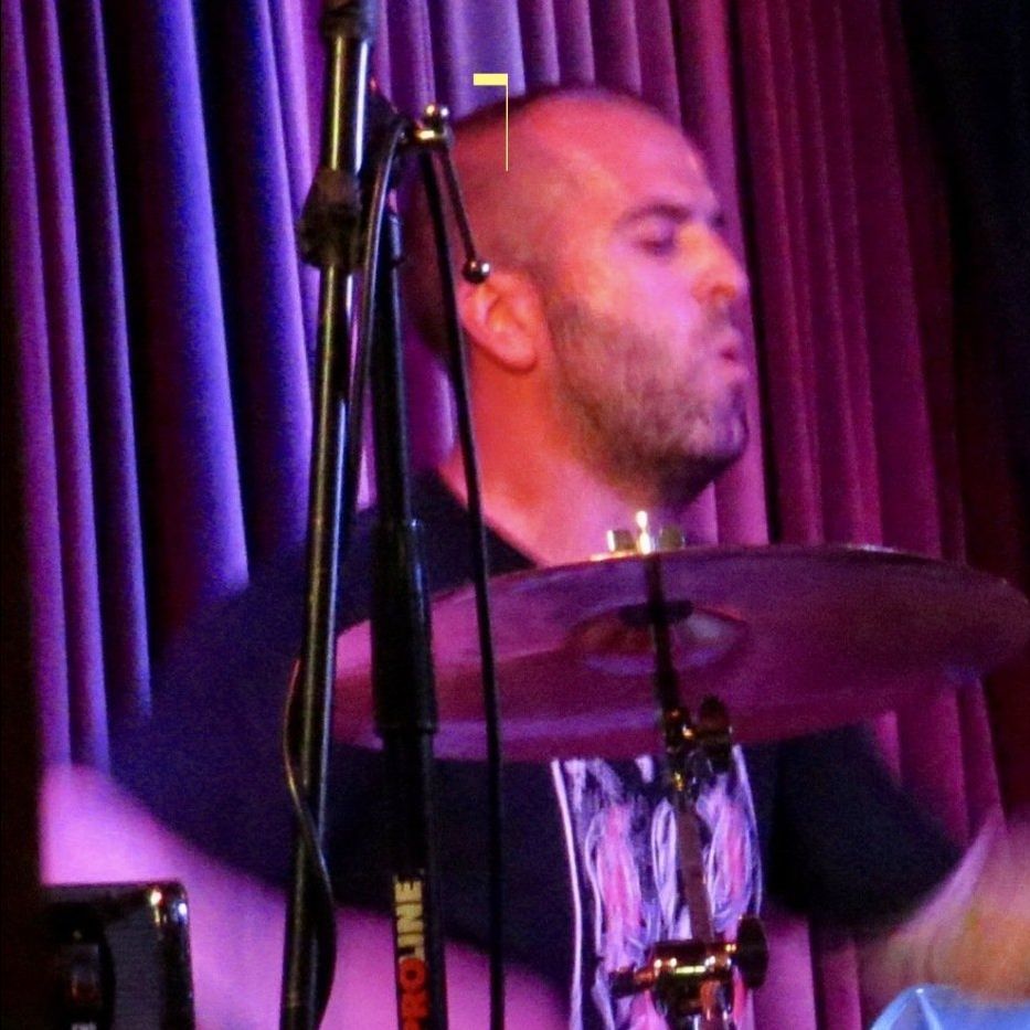 Drummer performing on stage behind a cymbal, with purple curtains and microphone stands