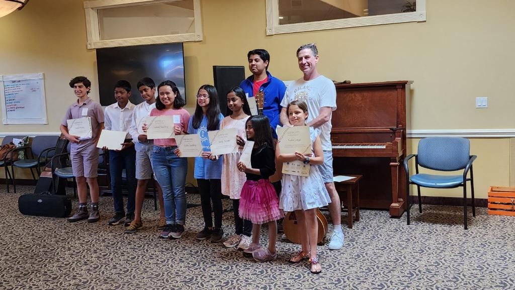 Group of children and adults posing indoors with certificates in front of a piano and chairs