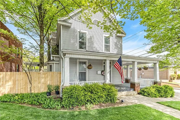Gray two-story house with white trim, front porch, American flag, and green lawn under leafy trees