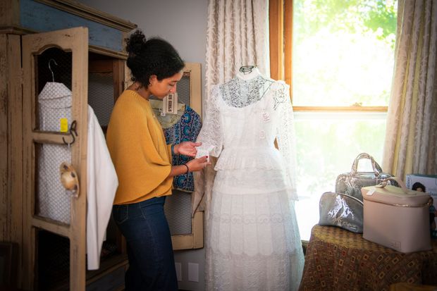 Person examining a white dress on a mannequin in a sunlit room with sewing supplies.