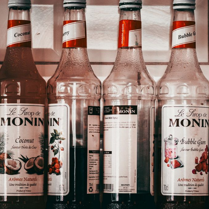 Four Monin syrup bottles: coconut, cherry, a blank label, and bubblegum. Red and white labels, clear bottles.