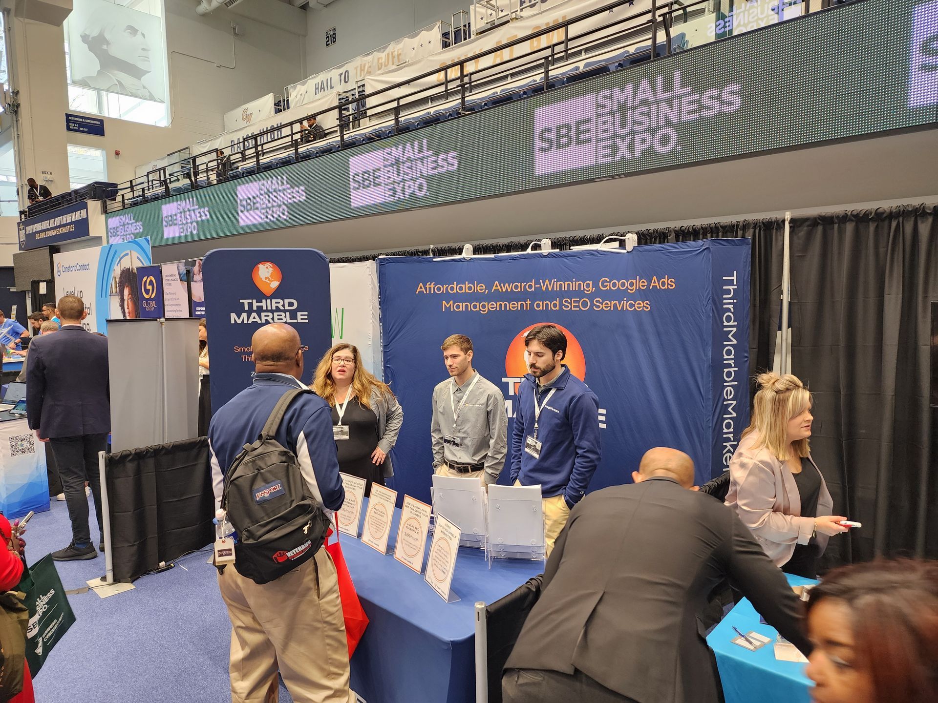 SEO Package: Conference booth with blue backdrop, people talking at tables, and a digital display overhead.