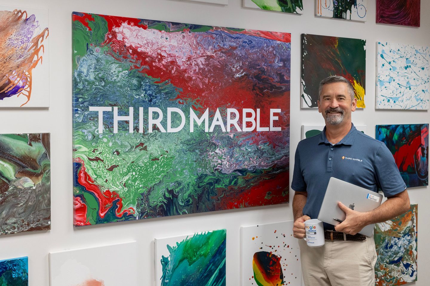 SEO Content Writing Service: Man holding a mug and folder beside colorful abstract art wall with “THIRD MARBLE” sign