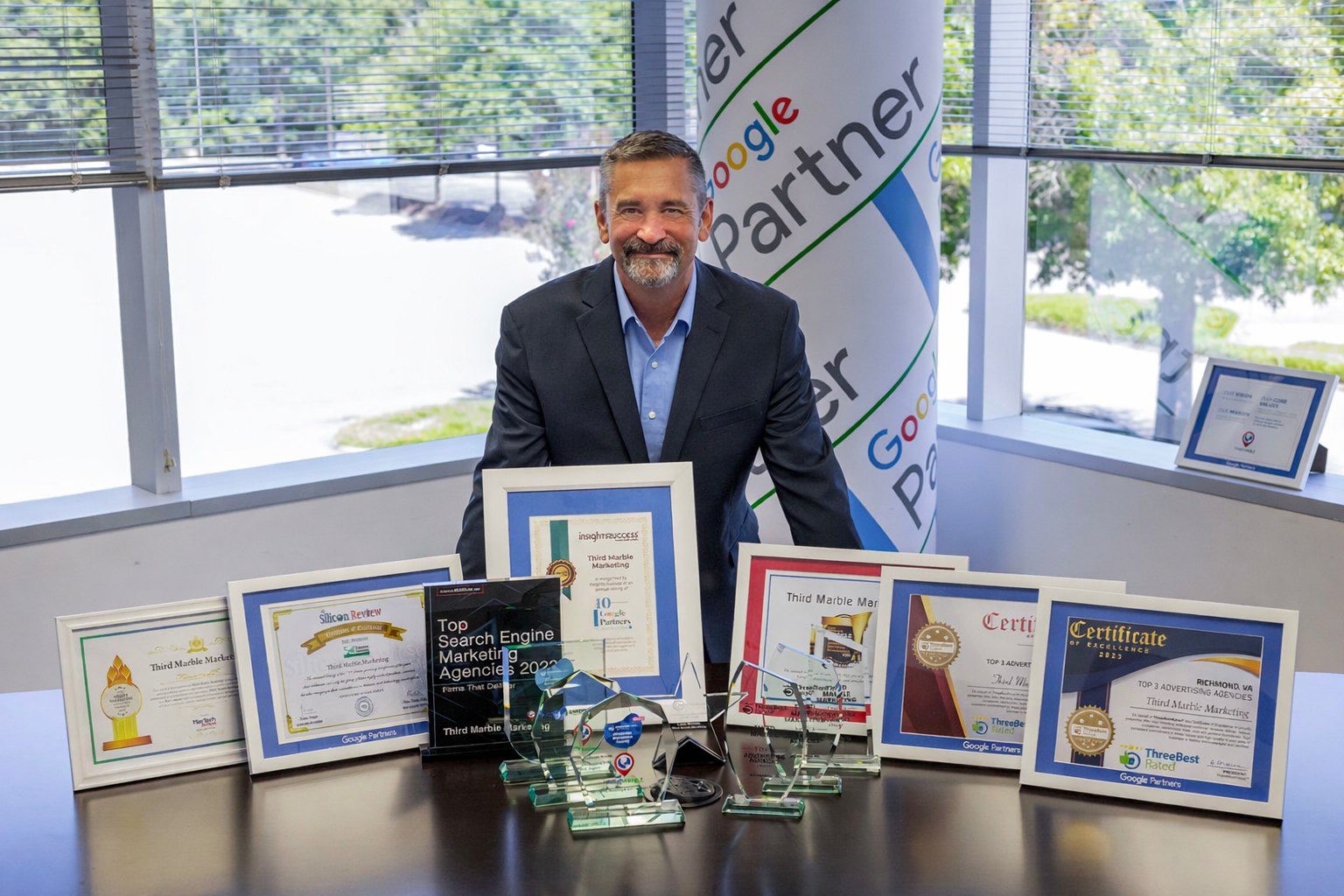 Google Ads Management Services: Man seated behind a table displaying framed certificates and awards in a bright office.
