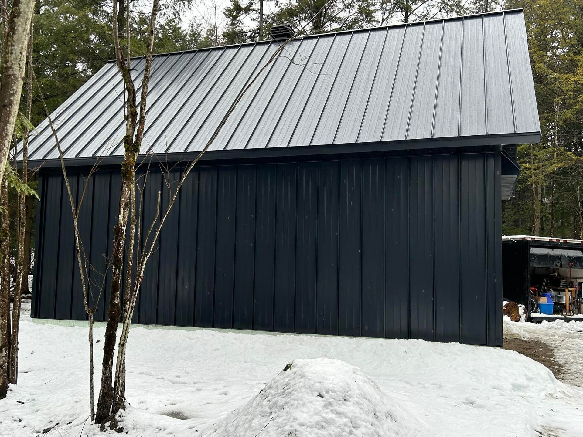 Un bâtiment noir avec un toit en métal est entouré de neige et d'arbres.