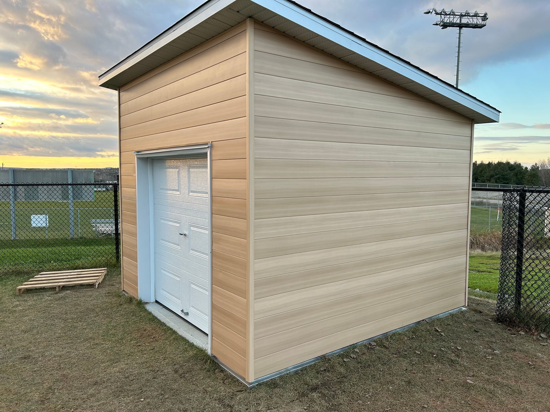Un petit hangar en bois avec une porte de garage blanche se trouve à côté d'une clôture grillagée.