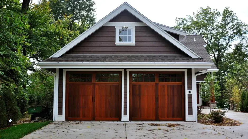 Two-car garage with dark wood doors and gray siding under a peaked roof, surrounded by trees Two-car garage with dark wood doors and gray siding under a peaked roof, surrounded by trees