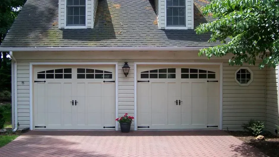 Two beige garage doors beneath dormer windows, with a brick driveway and flower pot in front Two beige garage doors beneath dormer windows, with a brick driveway and flower pot in front