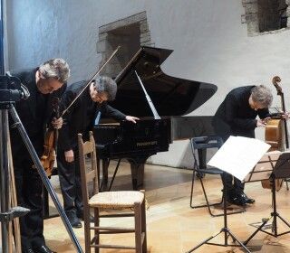 Tre uomini suonano violini e violoncello davanti a un pianoforte.