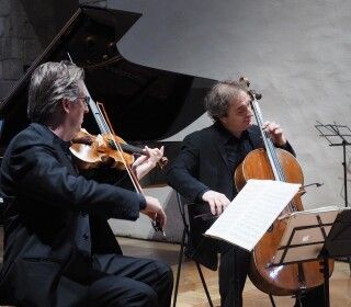 Due uomini suonano violini e violoncello davanti a un pianoforte.