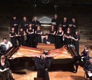 Un uomo è in piedi davanti a un grande pianoforte mentre un coro canta dietro di lui.