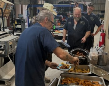 People serving food from trays at a kitchen counter in a busy cafeteria or restaurant. | Pete's Automotive