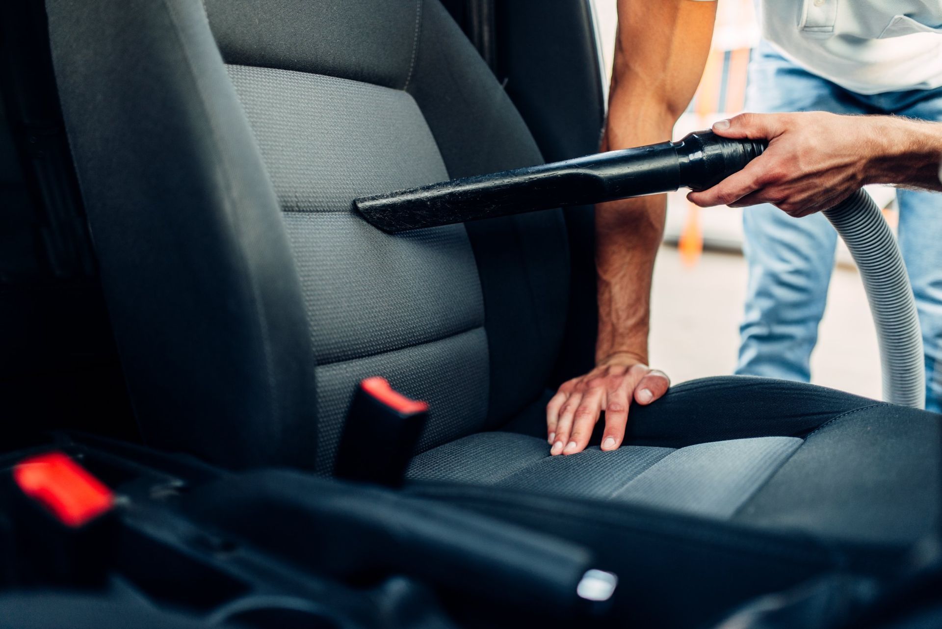 A person uses a vacuum attachment to clean the fabric upholstery of a car seat.