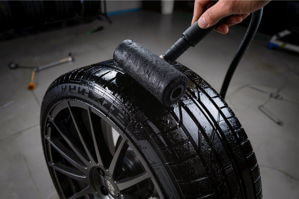 A hand uses a black foam roller to apply a glossy coating to the surface of a dark car tire in a workshop.