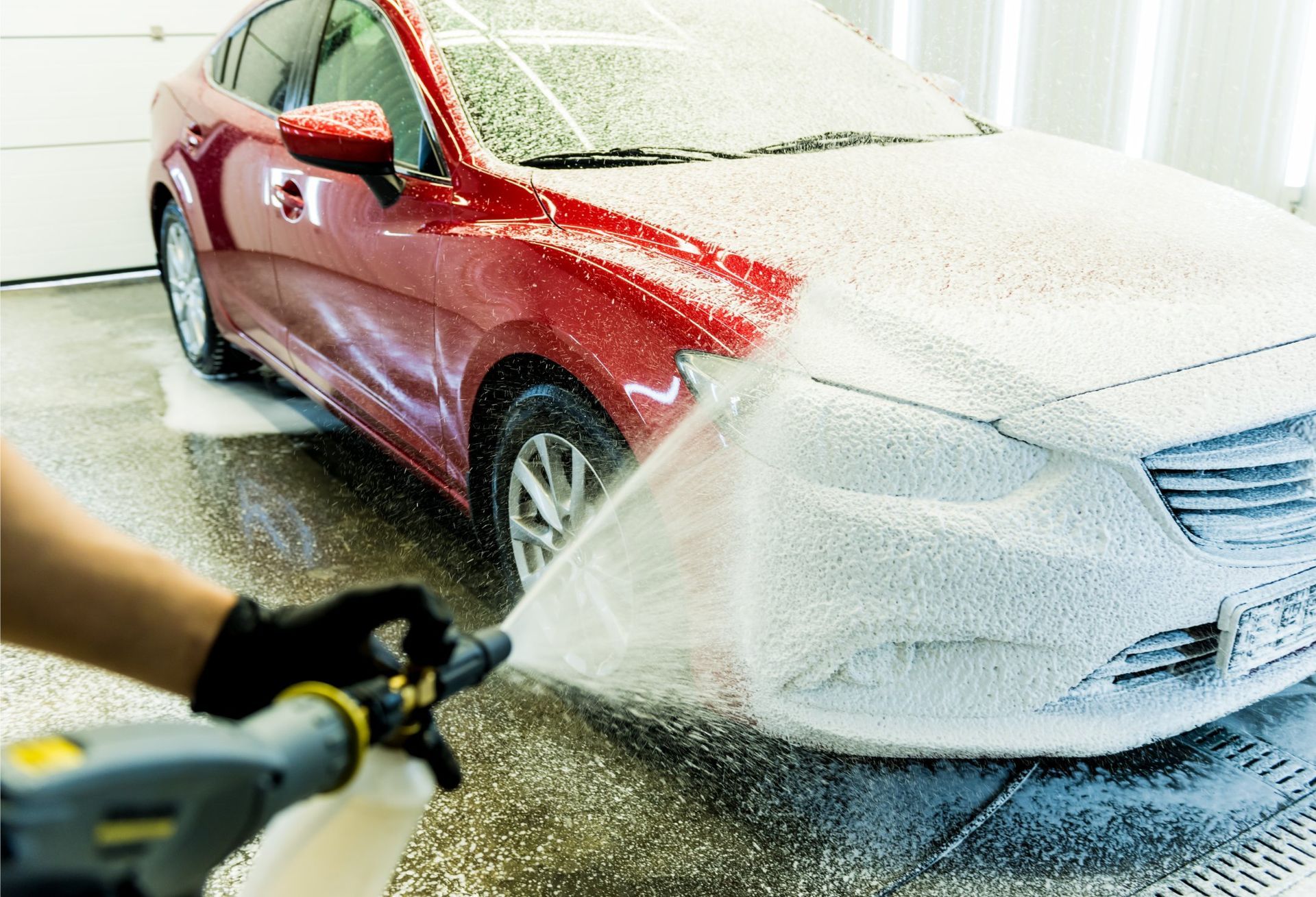 A hand wearing a black glove uses a foam sprayer to apply thick white soap onto a red car in a garage.