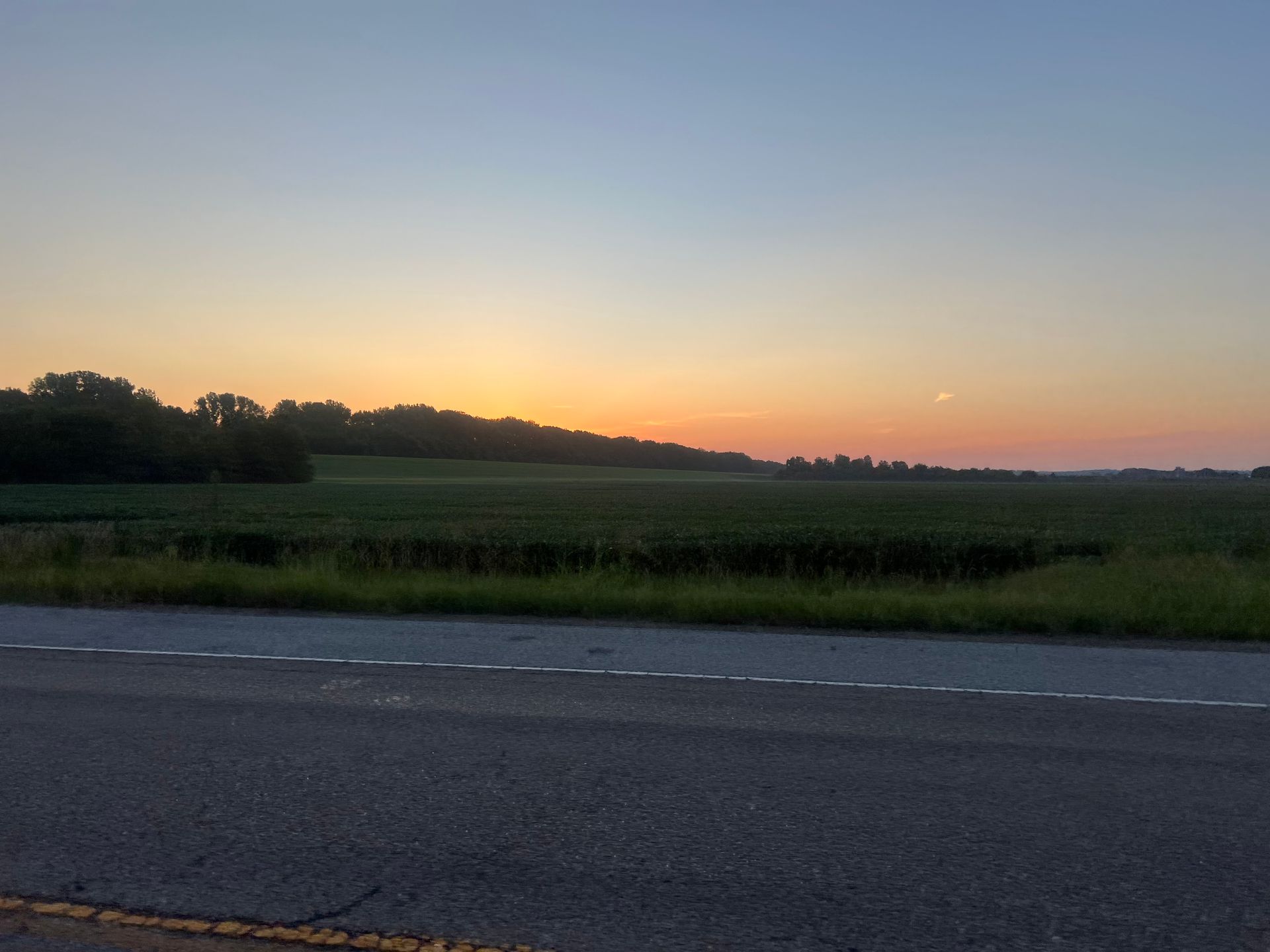Sunset over a green field and treeline, seen from a road. Orange, blue sky.