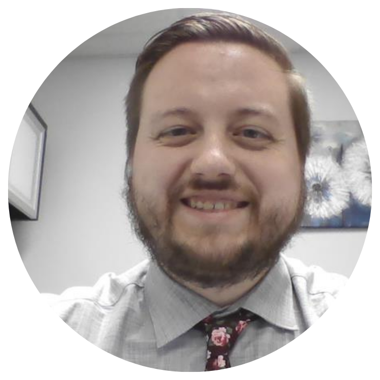Man with a beard and patterned tie smiles in a bright office setting.