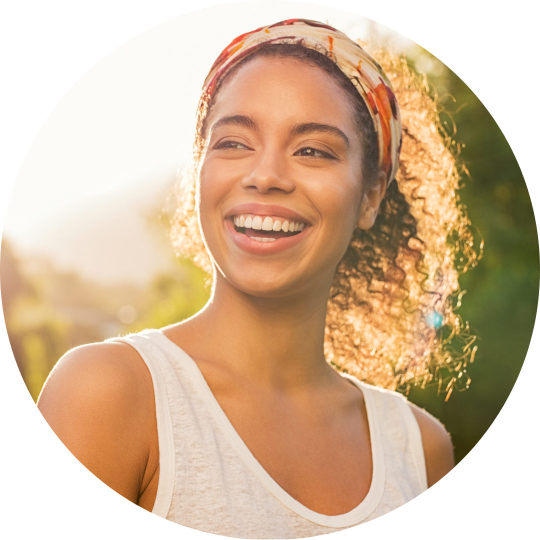 Woman with curly hair smiles, wearing a headband, tank top, and bathed in sunlight outdoors.