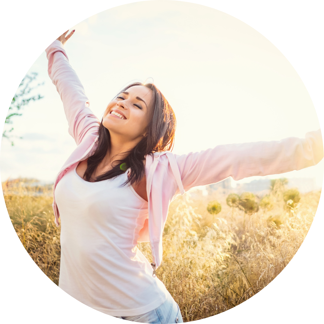 Woman with arms outstretched, smiling in a sunny field. She wears a white top and pink jacket.