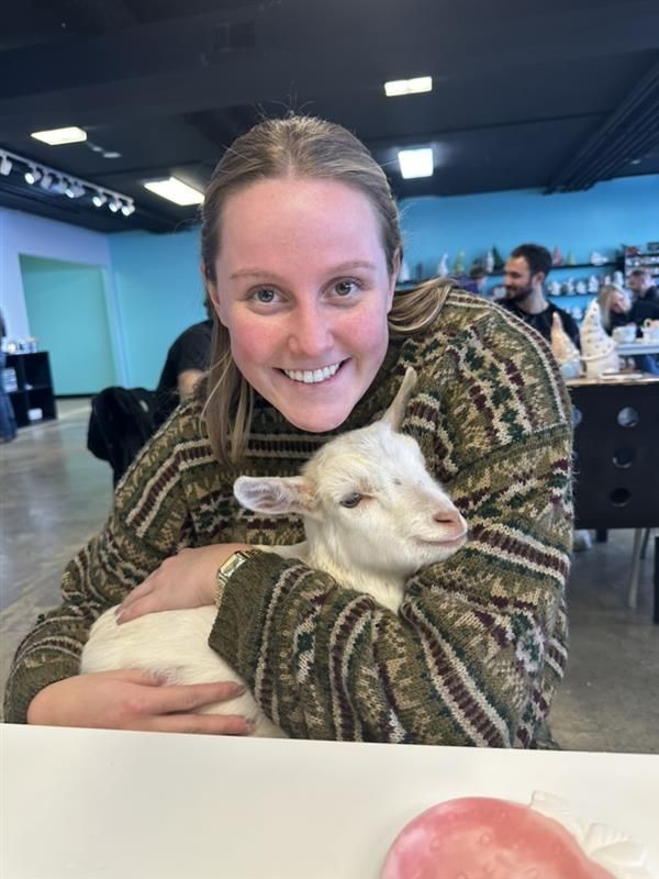 Woman in green sweater smiling, holding a white goat. Interior setting, smiling, looking at camera.