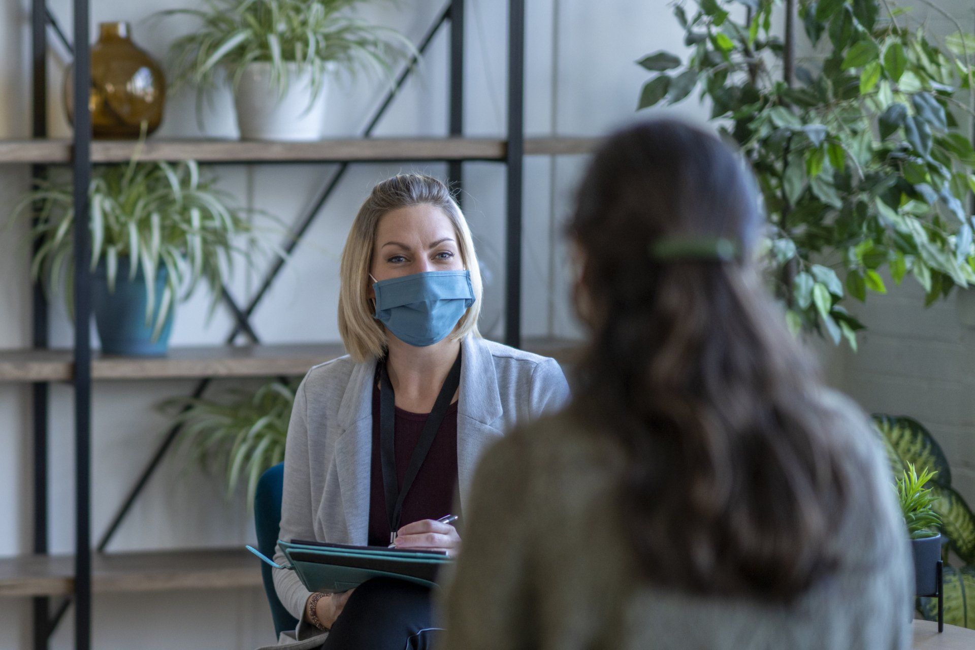 Woman in mask seated, facing another woman. Shelves of plants and decor in background.