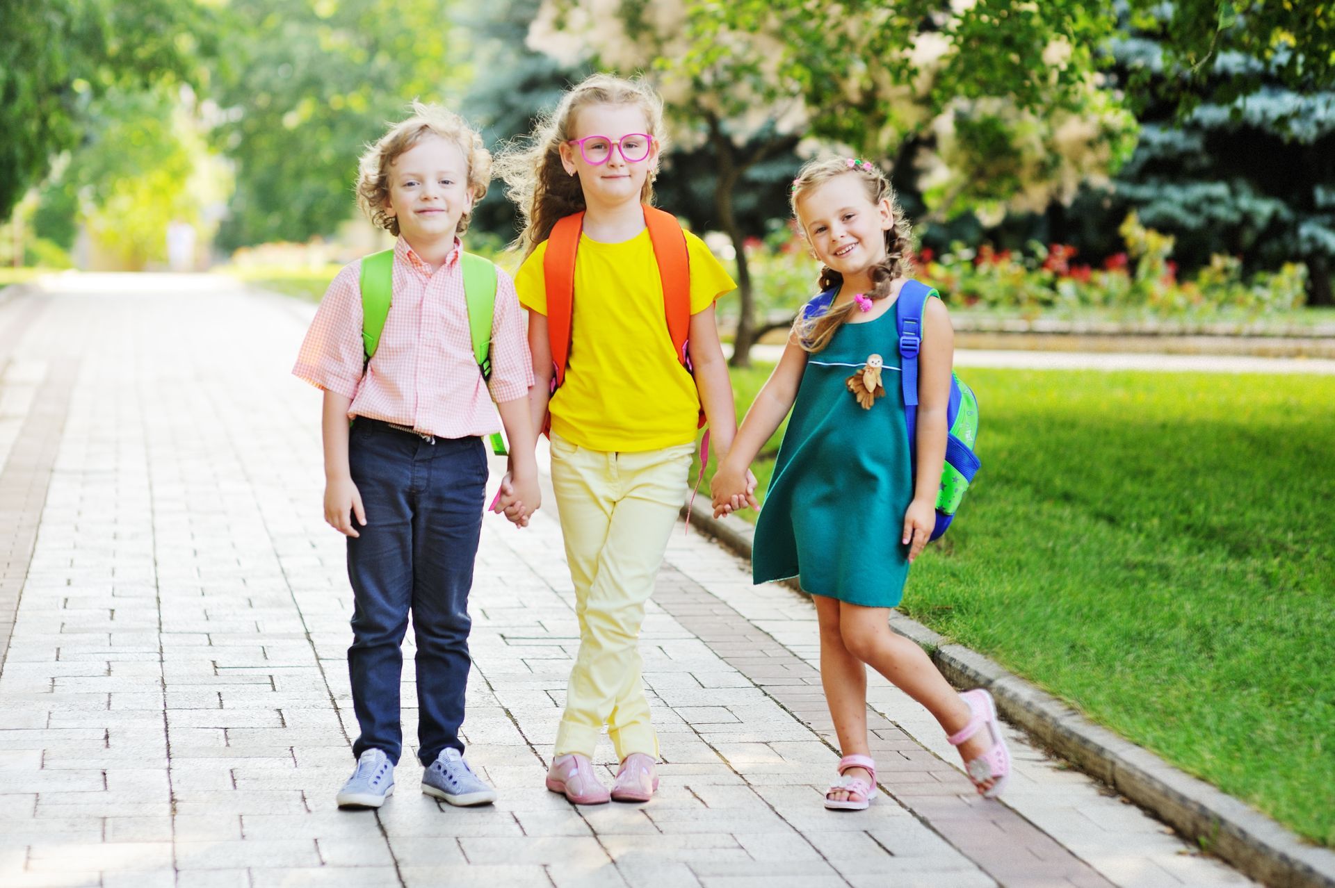 Three young children in colorful clothes and backpacks standing on a sidewalk outside, smiling and holding hands.