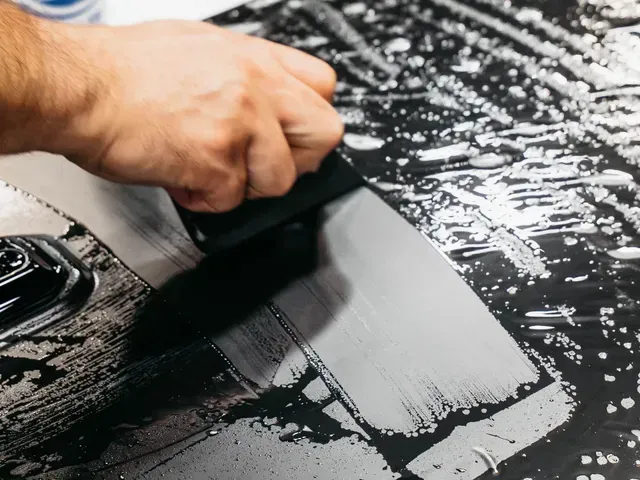 Hand spreading black liquid over a windshield with a squeegee, leaving a smooth gray streak