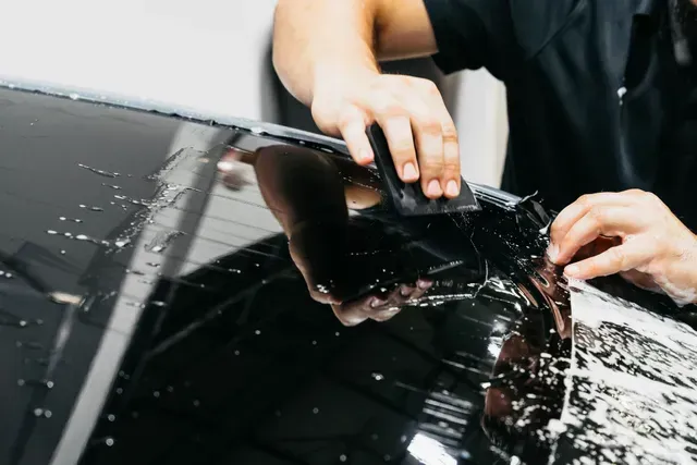 Hands applying clear film to a black car windshield, smoothing out bubbles and water droplets
