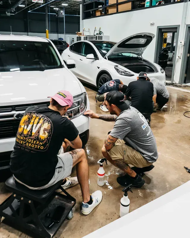 Three people crouch in a workshop detailing white cars, cleaning the front bumper area.