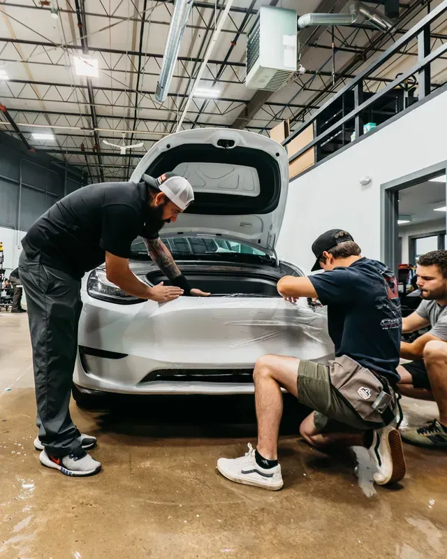 Three people inspecting the front of a white car in an auto shop, with the hood open.