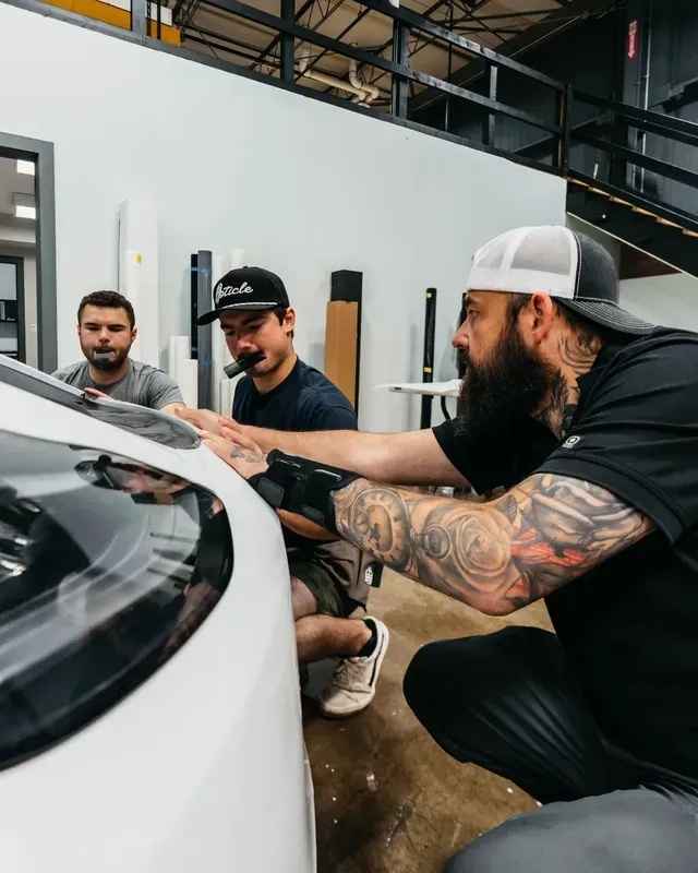 Three men in a workshop carefully inspect a white car’s windshield area.