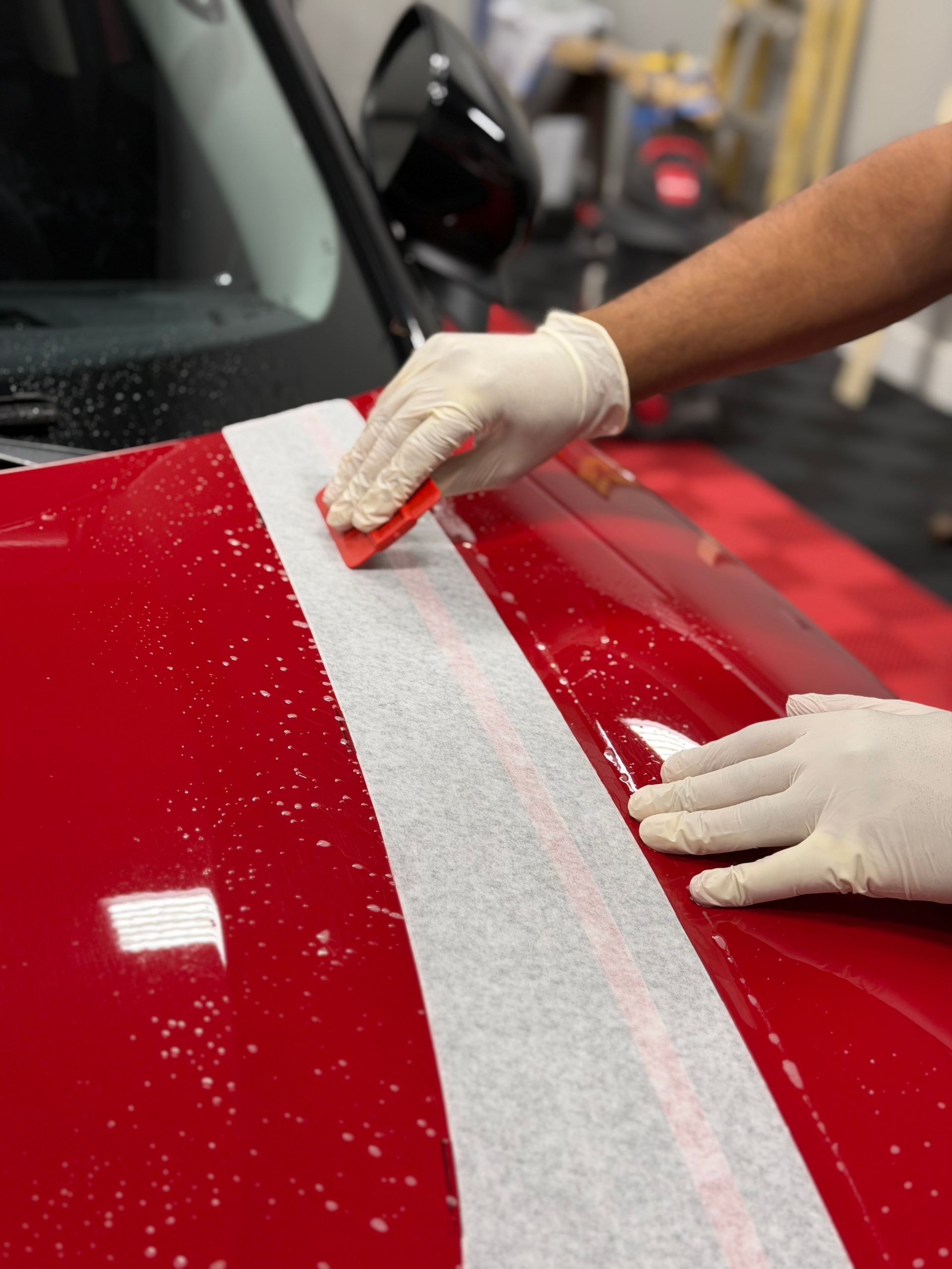 Hands apply a white racing stripe decal to a red car hood.