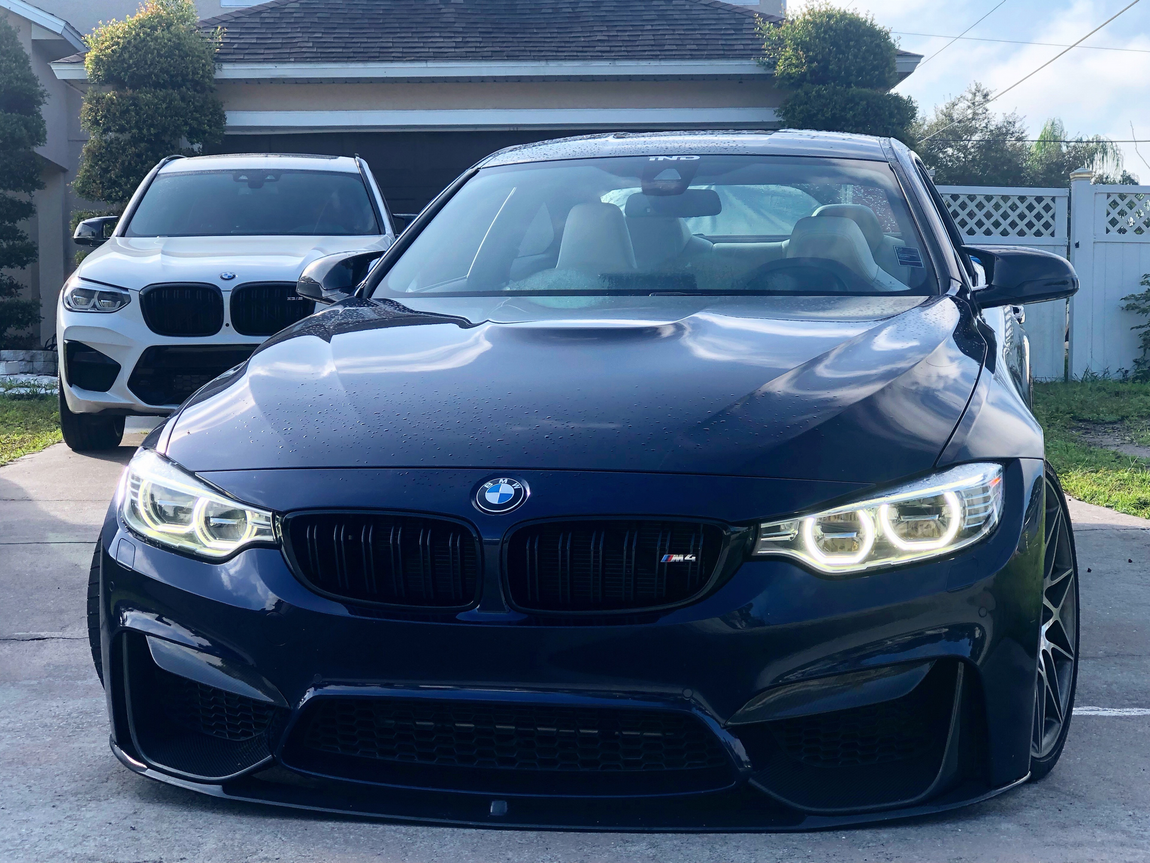 Blue BMW sedan parked in a driveway, front view, with a white SUV behind it.