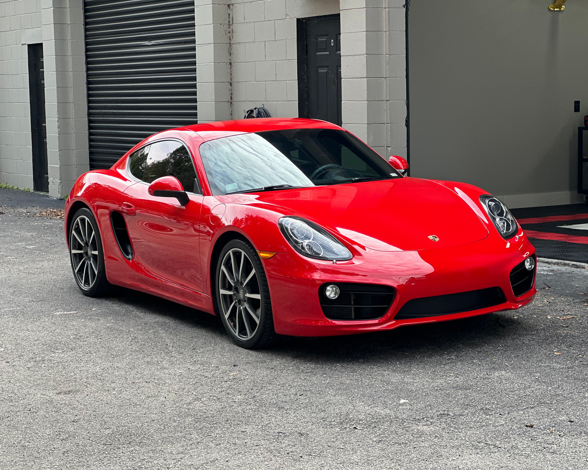 Red sports car parked on a gravel lot in front of a gray building with a garage door