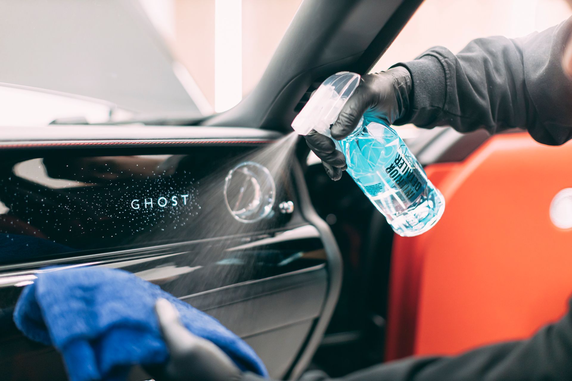 Gloved hand spraying cleaner on a car dashboard while wiping with a blue cloth