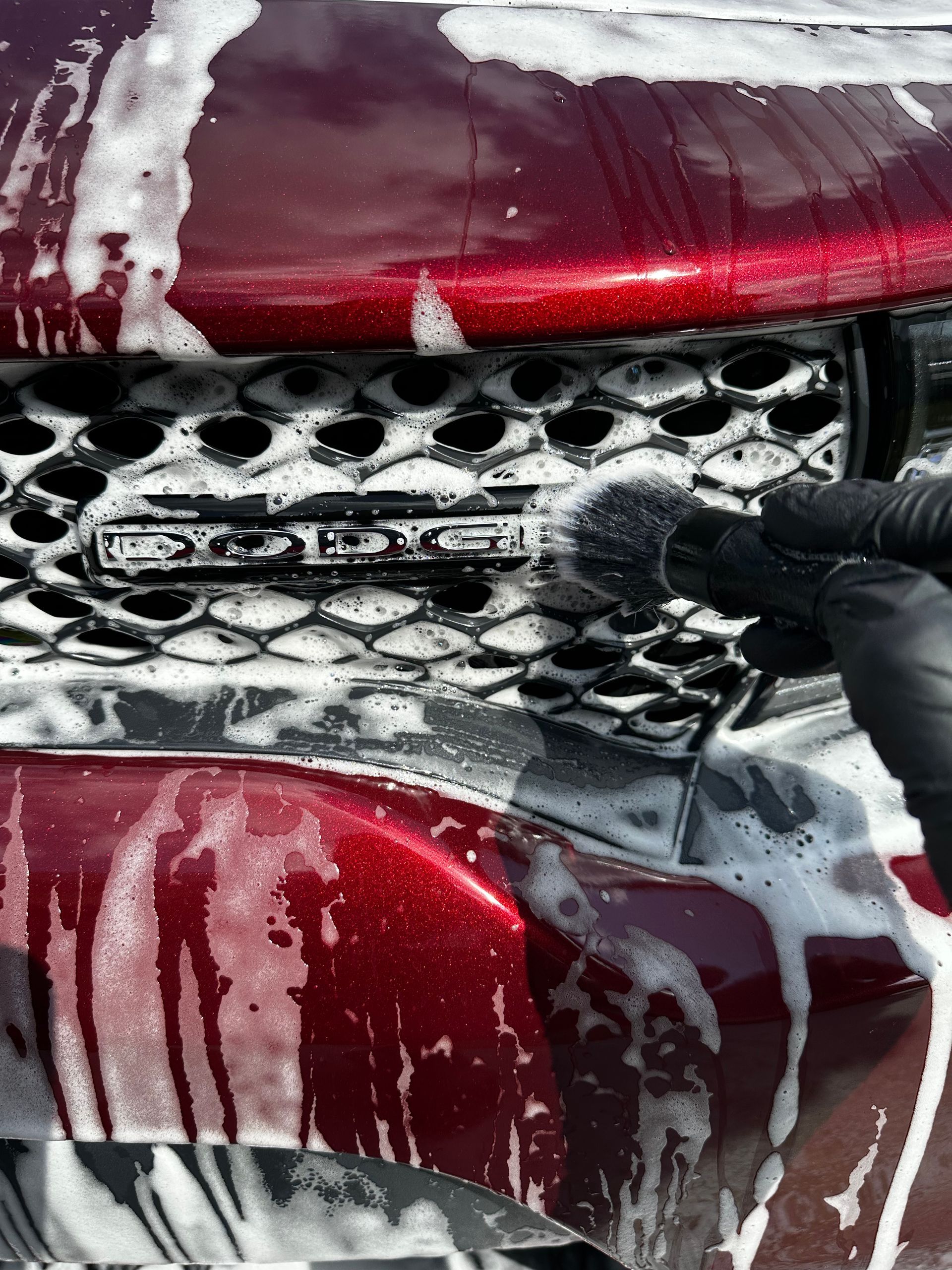 Close-up of a red car grille with white soap foam and a gloved hand washing it