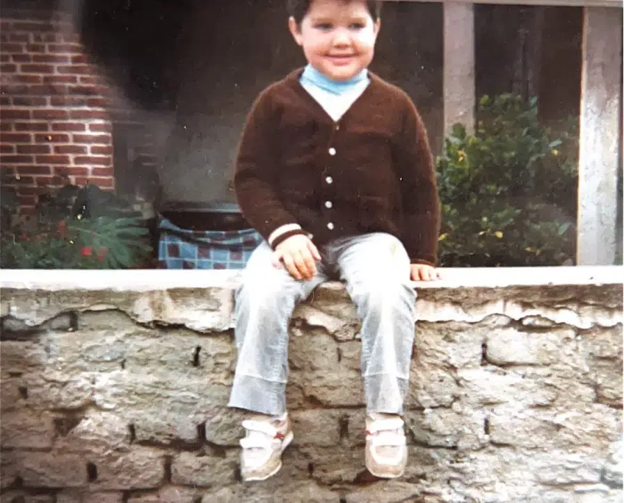 Un niño con un suéter marrón está sentado en un muro de piedra.