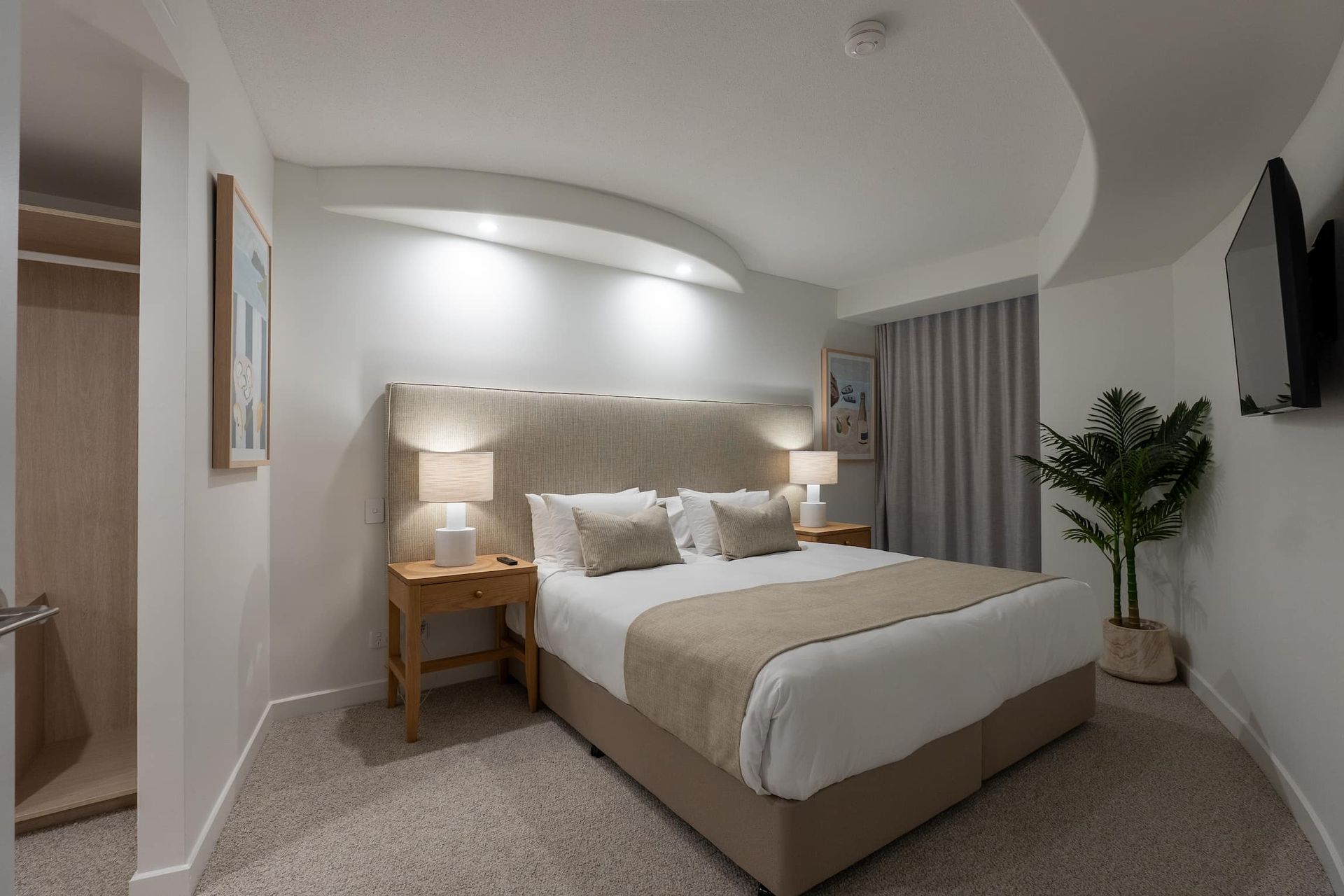 Modern bedroom with a queen bed, bedside lamp, neutral decor, and a potted plant.
