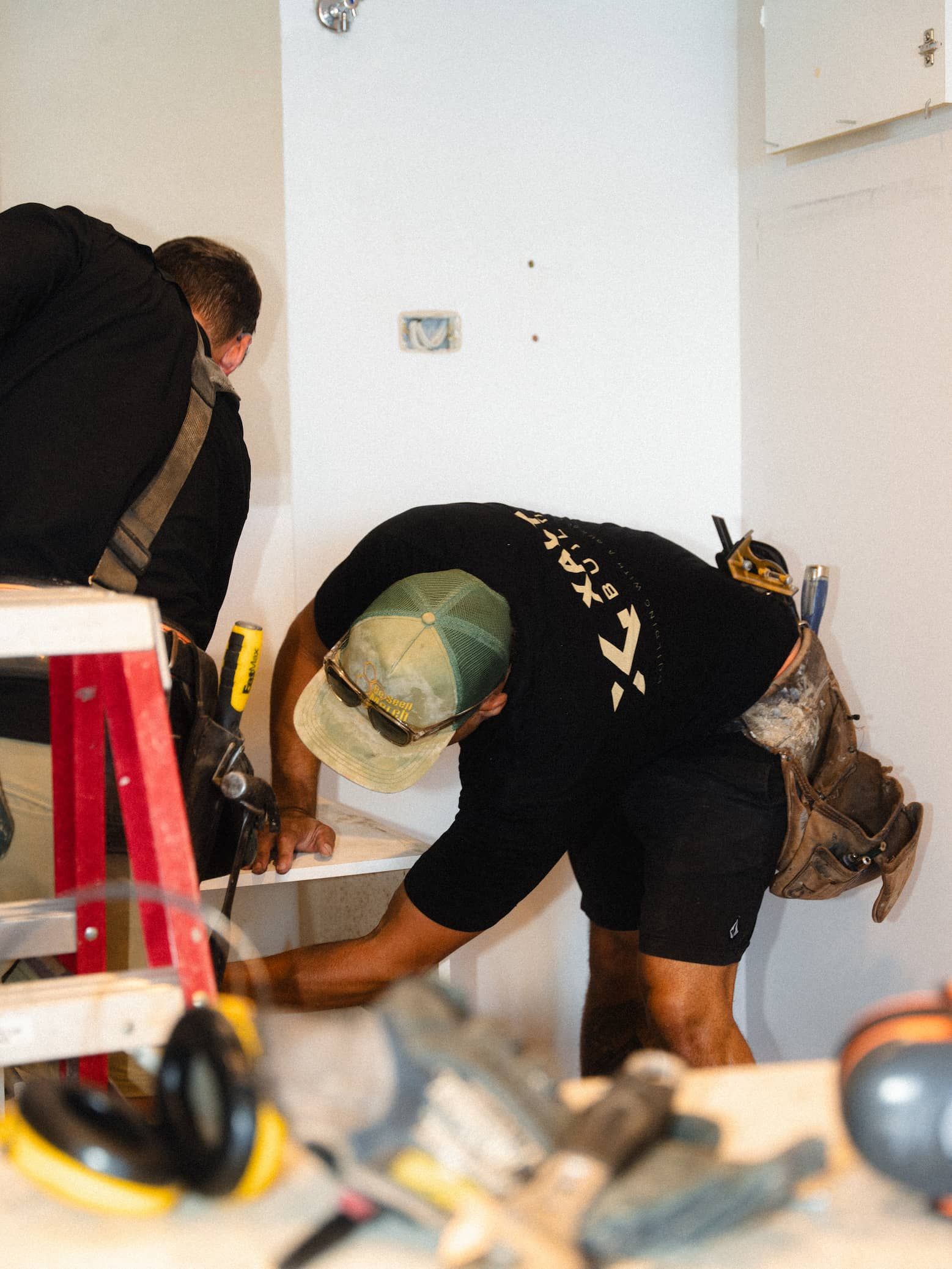 Two workers installing or repairing something inside a room, one bent over near a wall and another standing nearby.