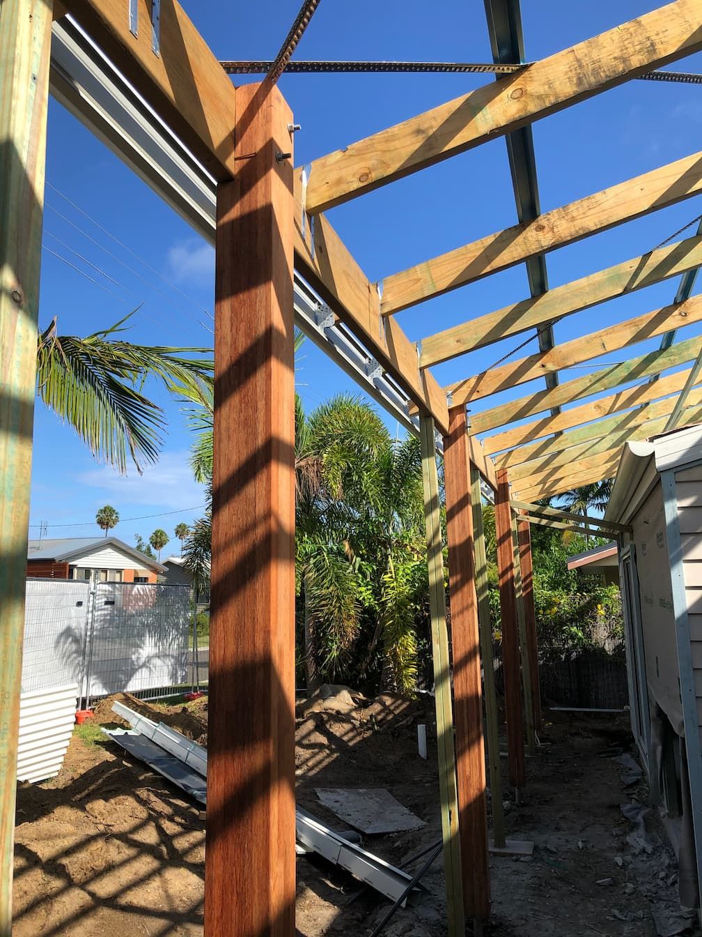 Partially built wooden pergola with vertical posts and roof beams beside a narrow outdoor walkway under blue sky