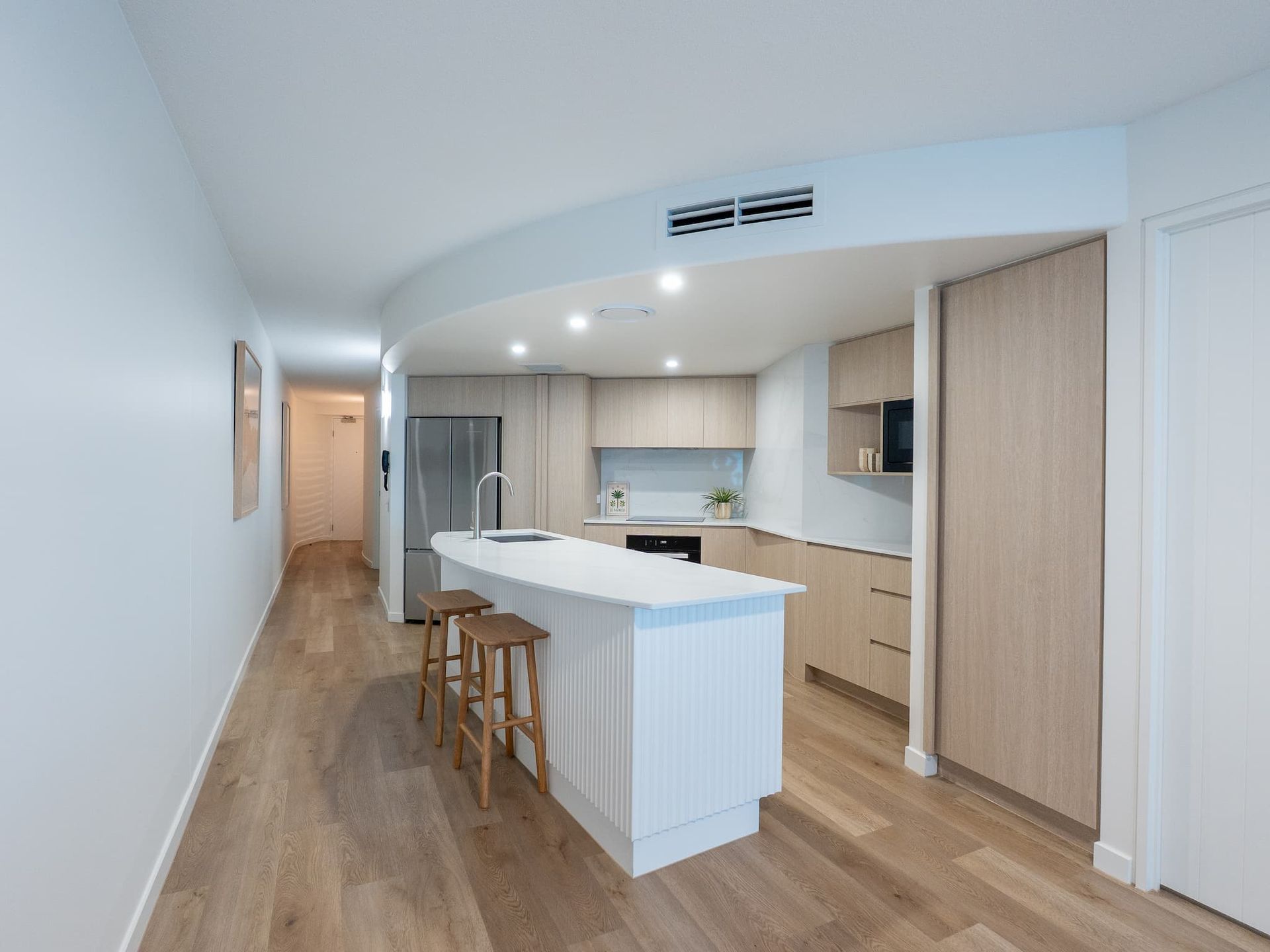 Bright modern kitchen with white island, wood cabinets, and stools in a long open-plan room