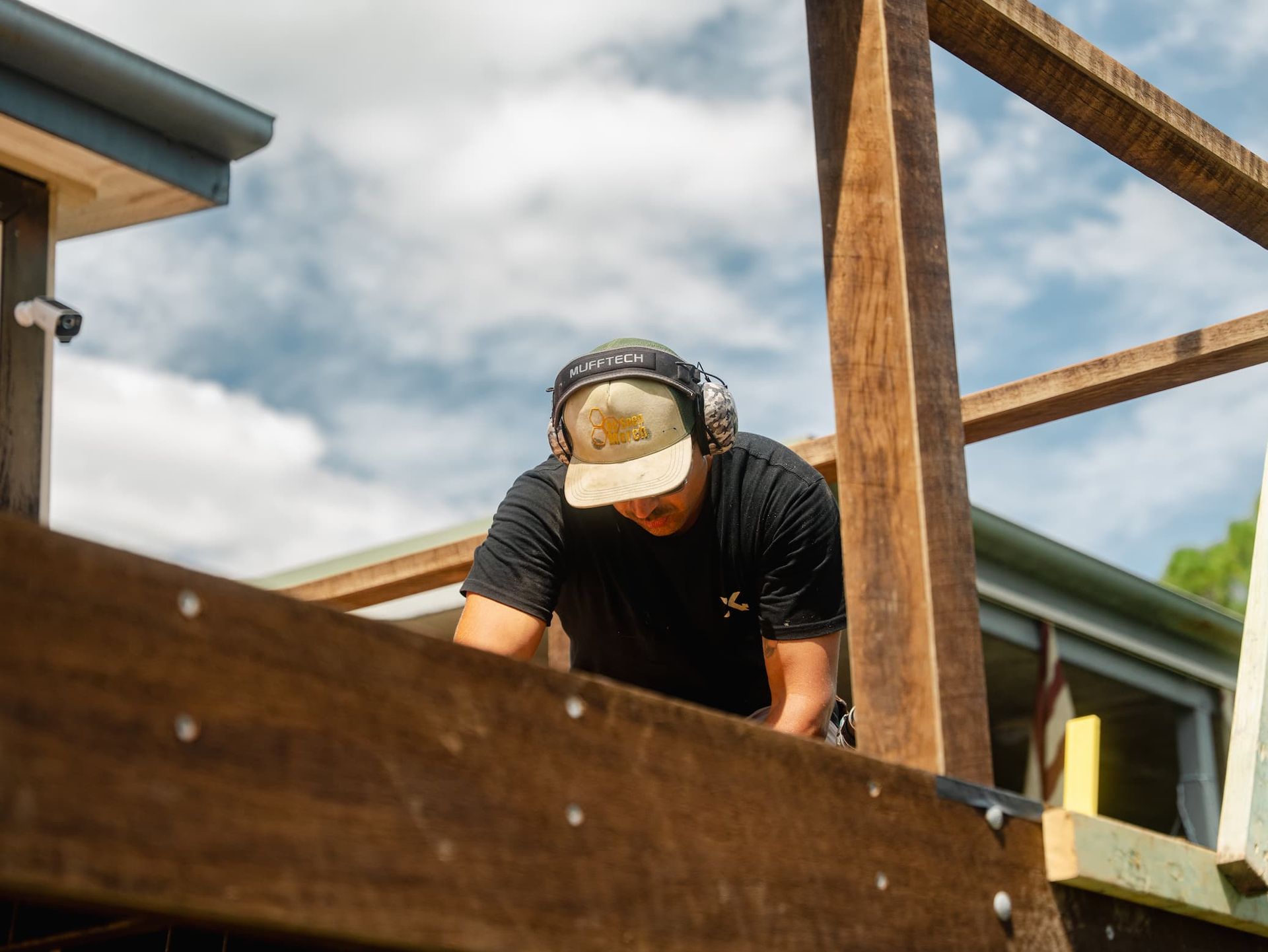 Construction worker wearing a hard hat and earmuffs leans over a wooden frame on a house build site.