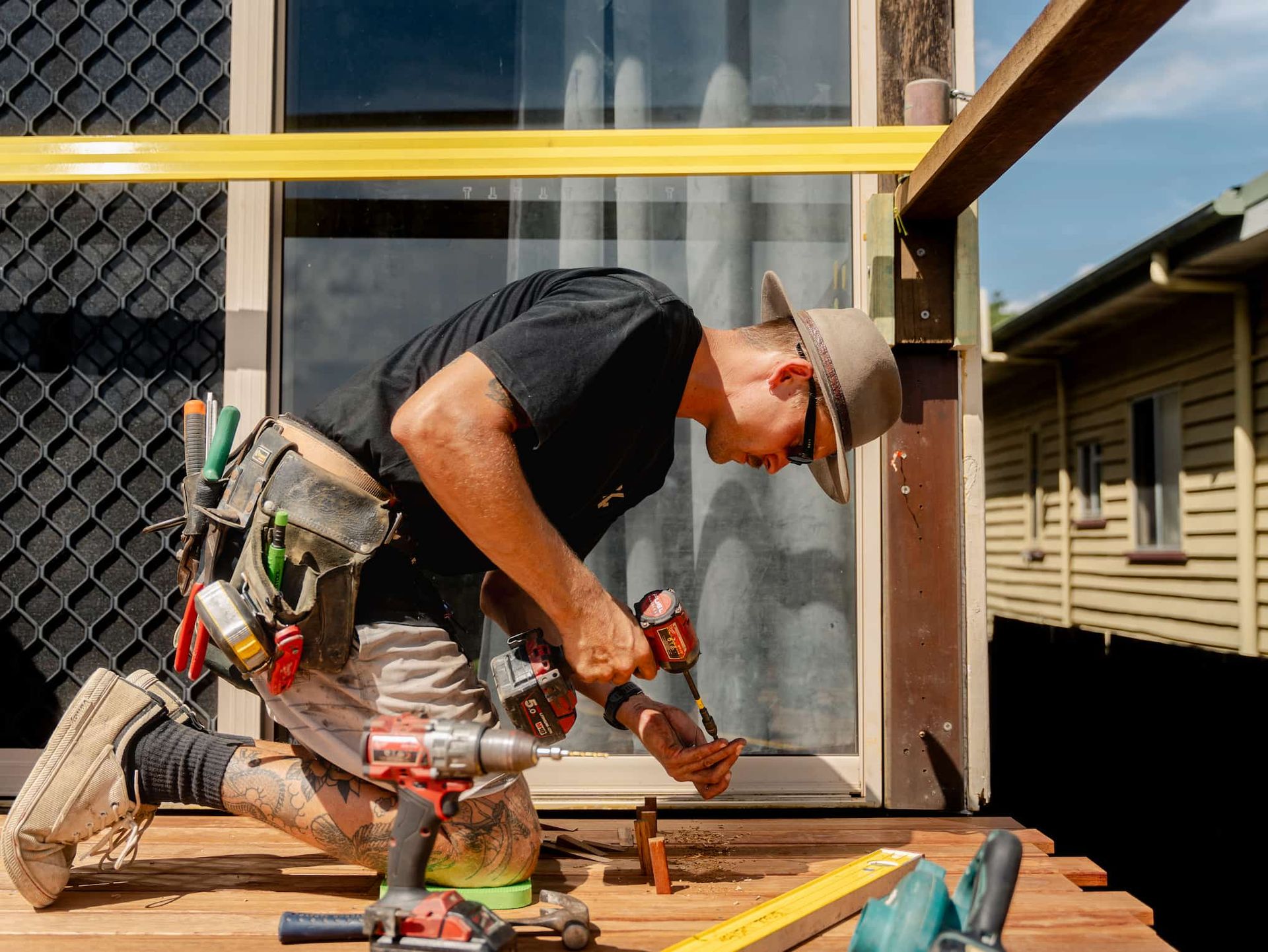 Worker using power tools while kneeling on a wooden deck beside a house under construction