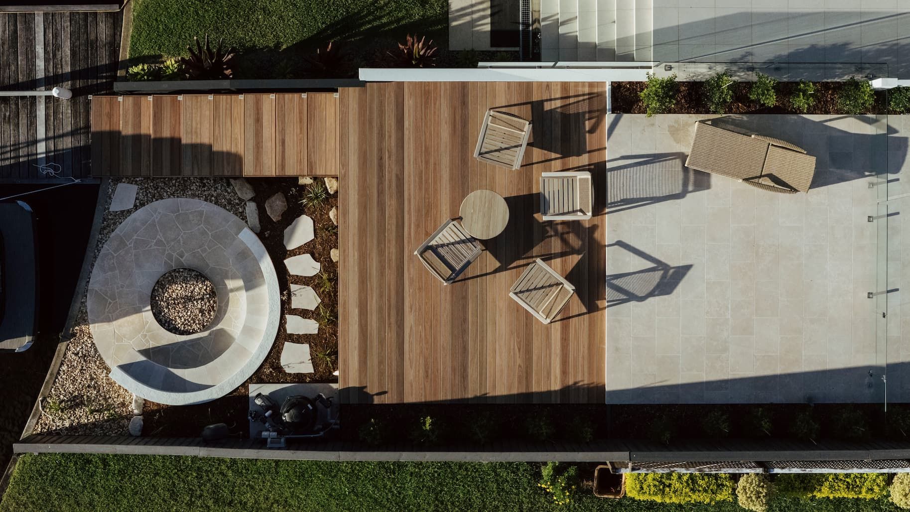 Overhead view of a wooden patio with a circular fire pit, chairs, and an outdoor dining table next to paving stones