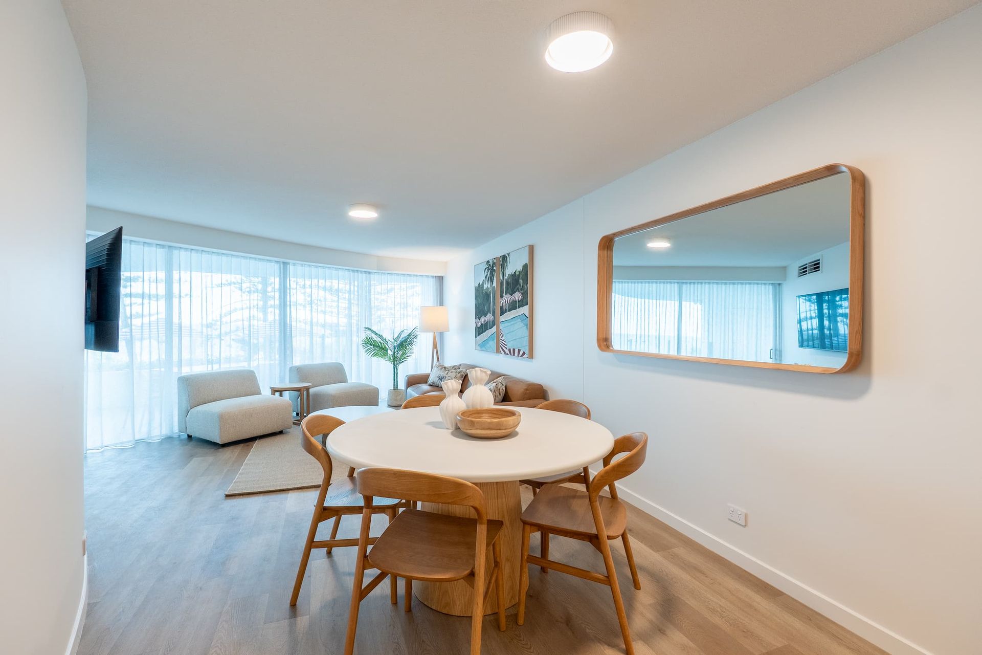 Bright dining and living room with round table, wooden chairs, sofa, and large window with sheer curtains