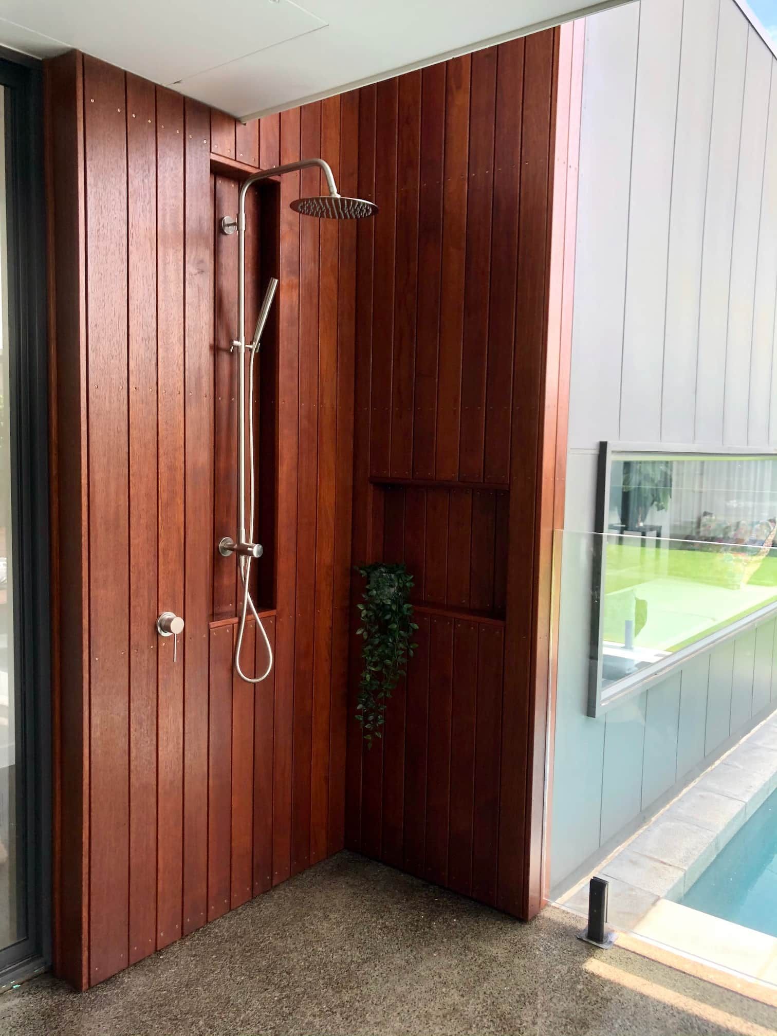 Outdoor shower with wood-paneled walls, stainless fixtures, and an open view to a pool area.